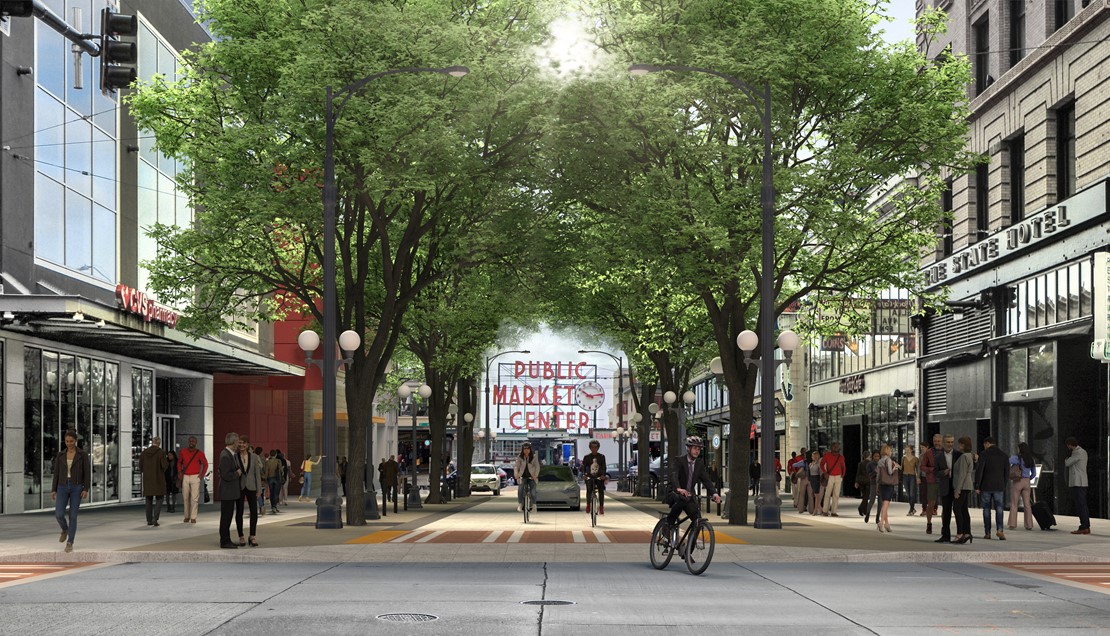 Wide sidewalks and a curbless street on the 100 Pike Street block filled with pedestrians. The street is lined with trees on both sides and people riding bicycles alongside cars with the Public Market sign in the background